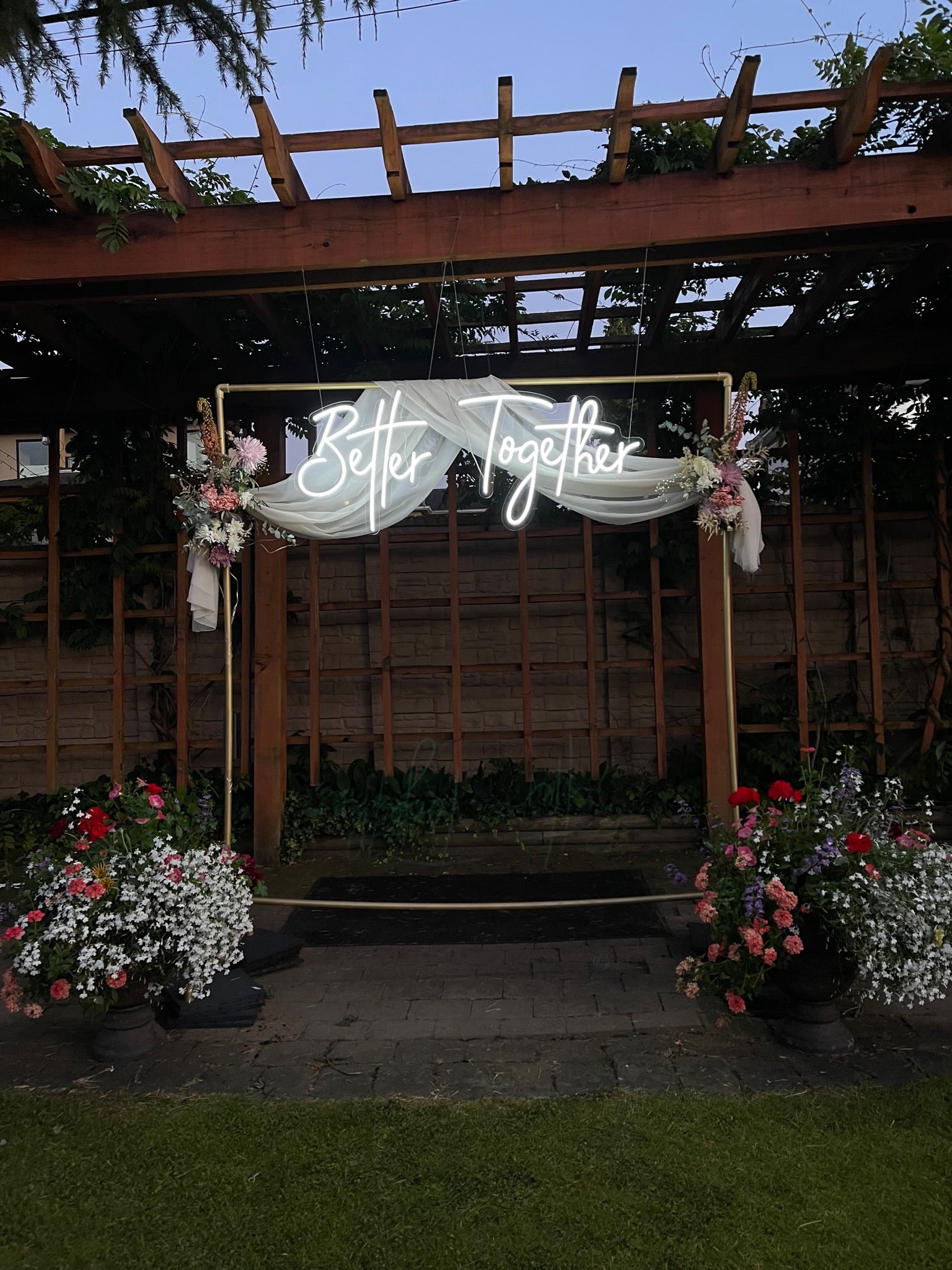 Decorative arch with 'Better Together' sign and floral arrangements in a garden setting, neon sign for wedding