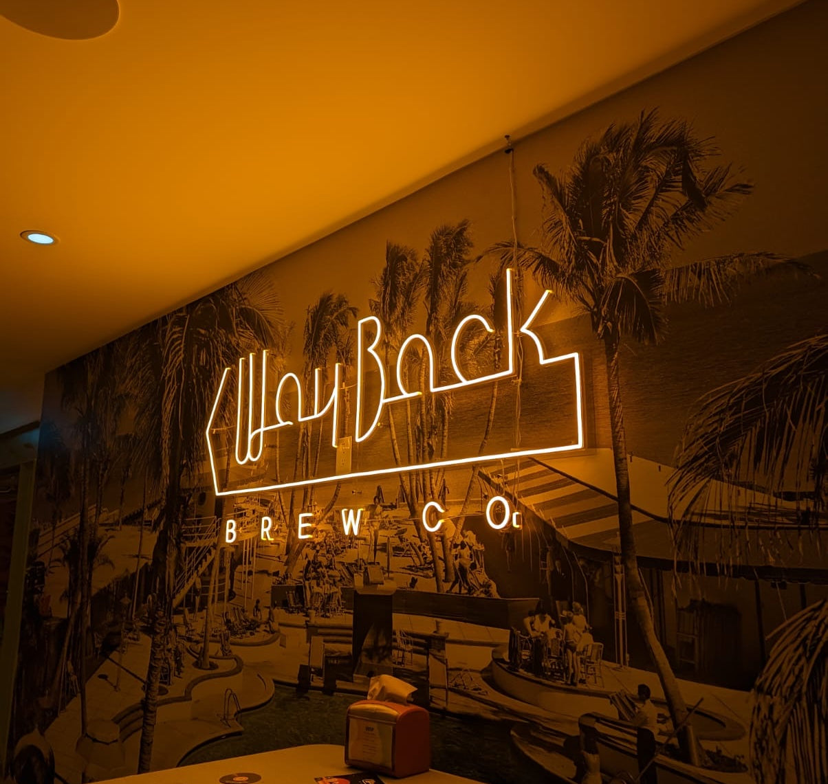 Neon sign for Wayback Brew Co. in a dimly lit room with palm tree mural.